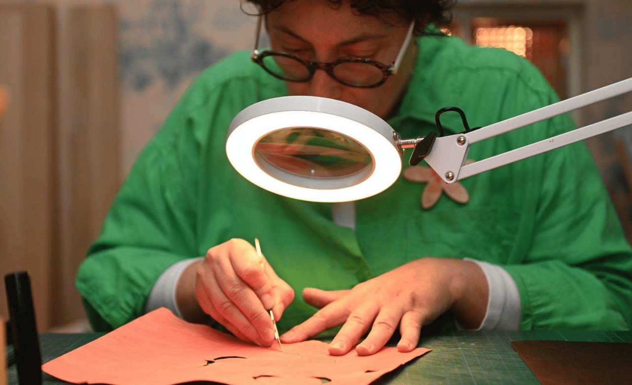 Une artisane d'art en pleine réalisation d'un motif en marqueterie cloisonnée sous une loupe éclairée, illustrant la fabrication artisanale française sur mesure et l'attention portée aux détails. Atelier Nathalie Melon